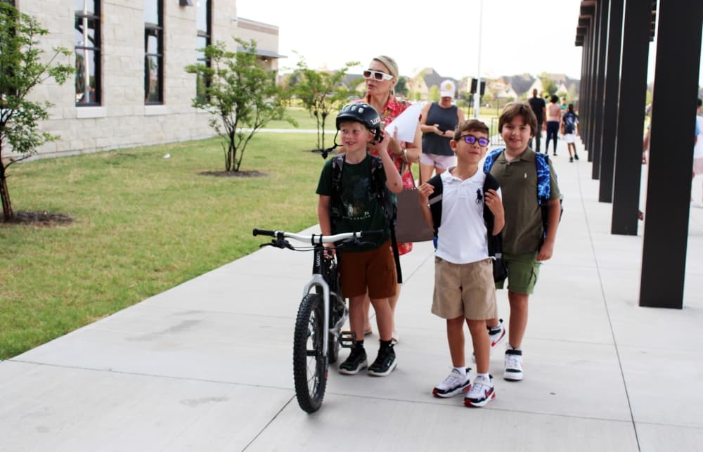 Three boys arriving at school. One boy is pushing a bike. 