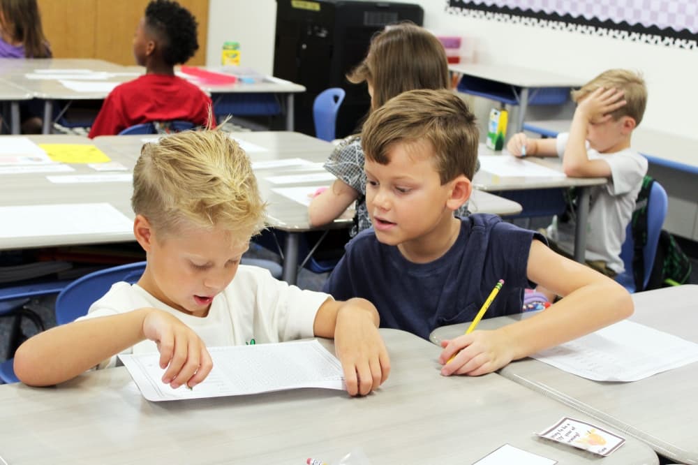 Two elementary aged males working writing on paper while seated at desks. 