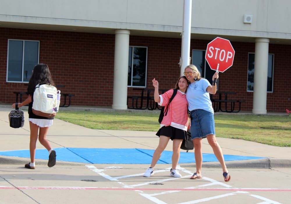 An adult female, holding a crossing guard stop sign, gives a side hug to a teen female.
