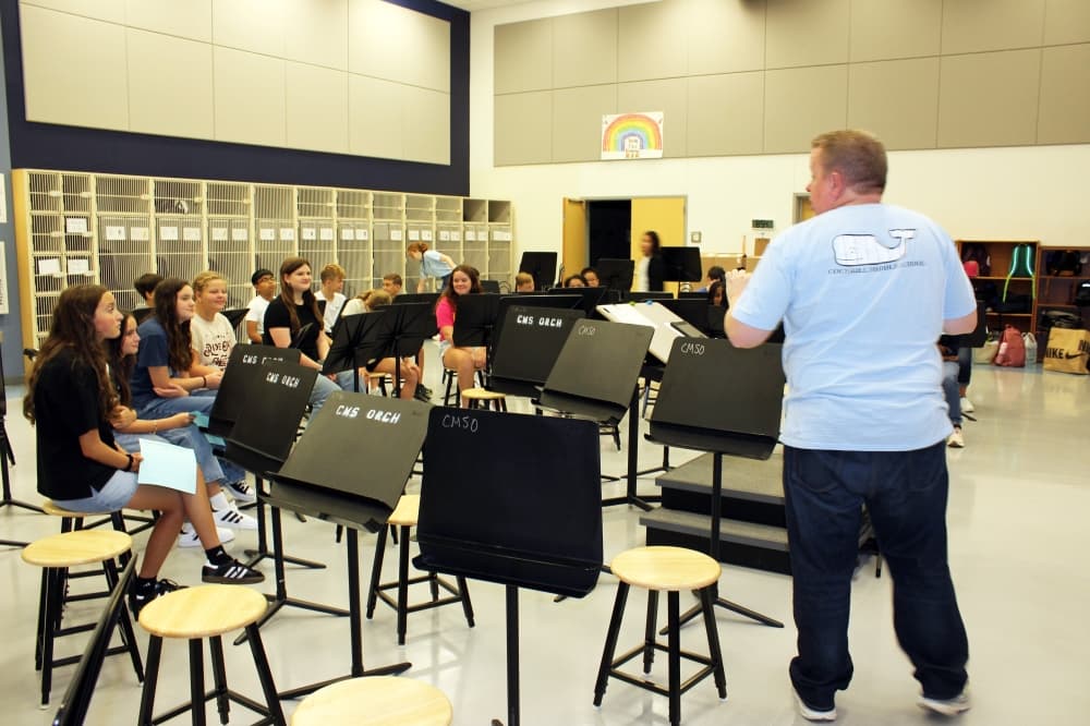 Adult male standing in front of students seated behind music stands. 