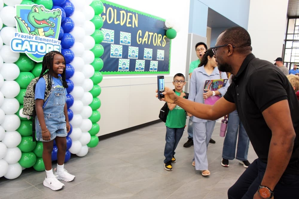 Adult male taking a phone photo of a child in front of a multi-colored balloon photo prop. 