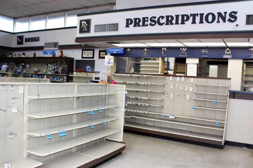 Interior of a store with empty shelves and a sign over a counter reading Prescriptions. 