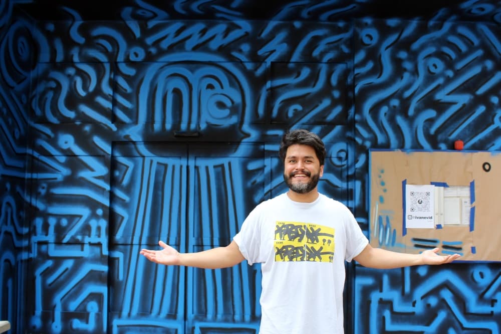 Male standing with arms outstretched to each side while standing in front of a blue and black mural. 