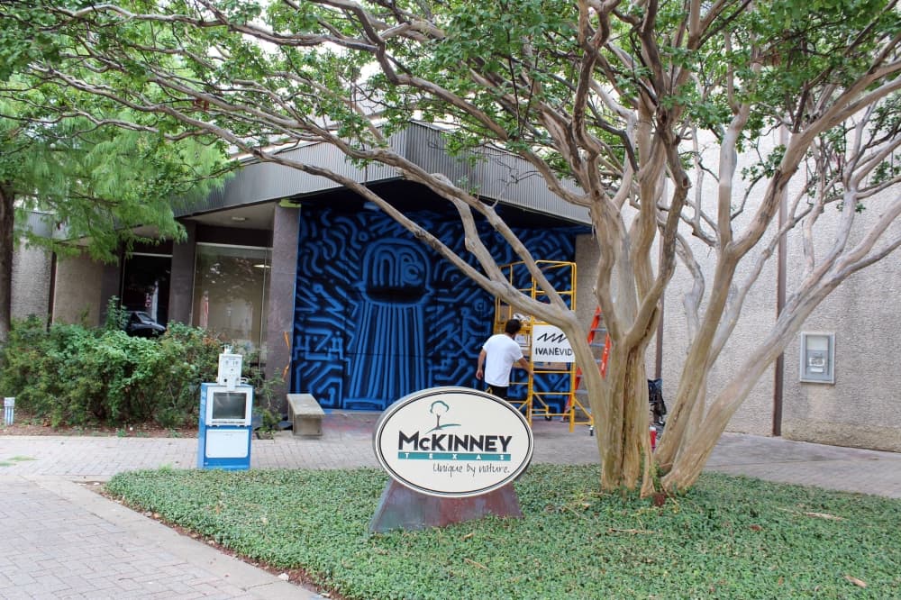 Exterior of a building with a blue and black mural and a small sign featuring the city of McKinney logo. 