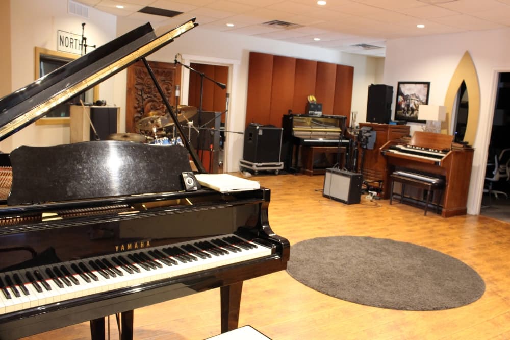 Room with a black baby grand piano, drums and upright pianos. 
