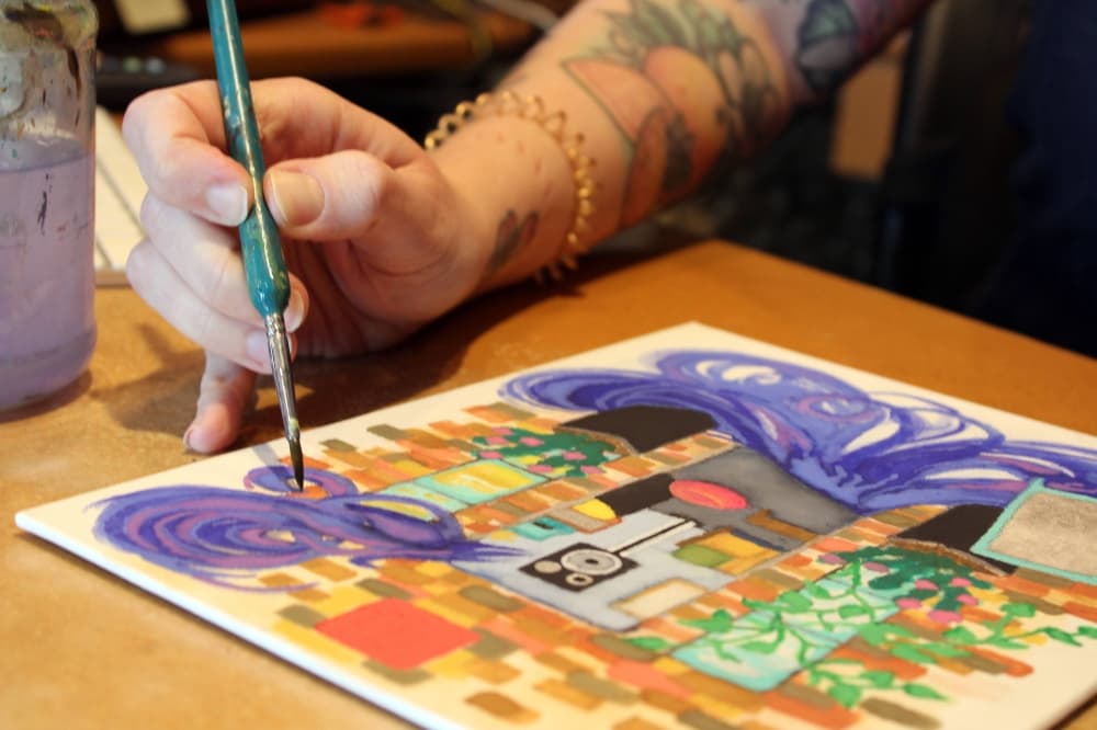 Person's hand painting a multi-colored scene on paper. 