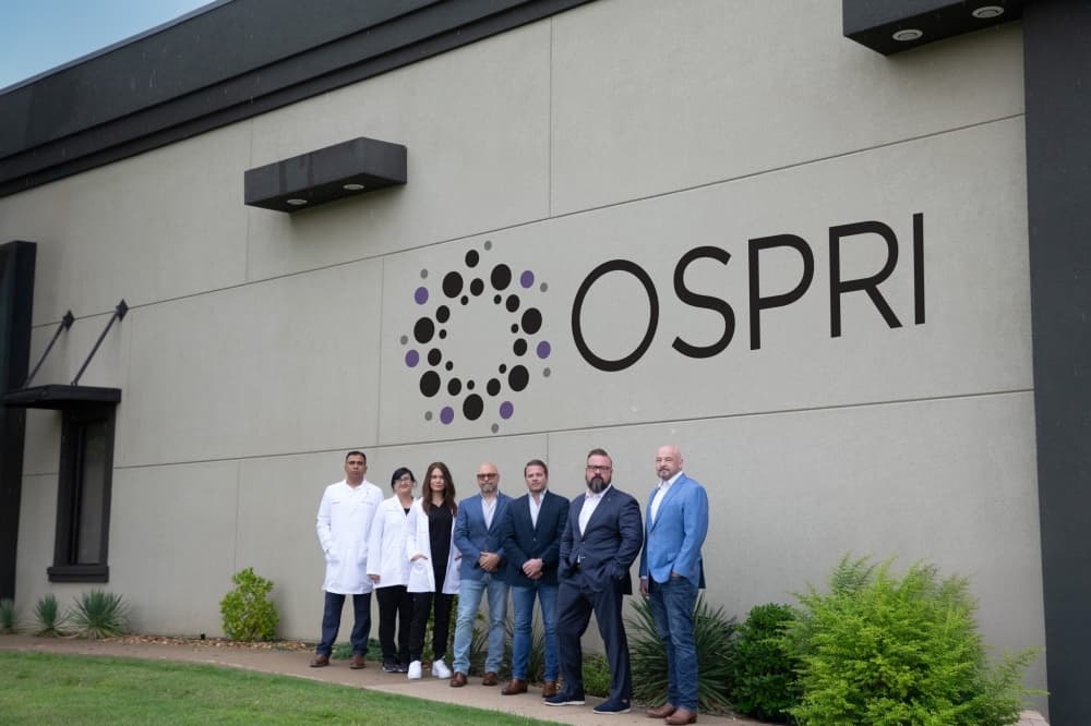 OSPRI officials pose under a company sign in McKinney. From left: Dr. Rahul Sharma, Dr. Sadia Almas, Dr. Alaina Vincent, Dr. Rob Carpenter, cofounder Jeremy Adamic, cofounder Steve Storey, cofounder Randy Phelps.
