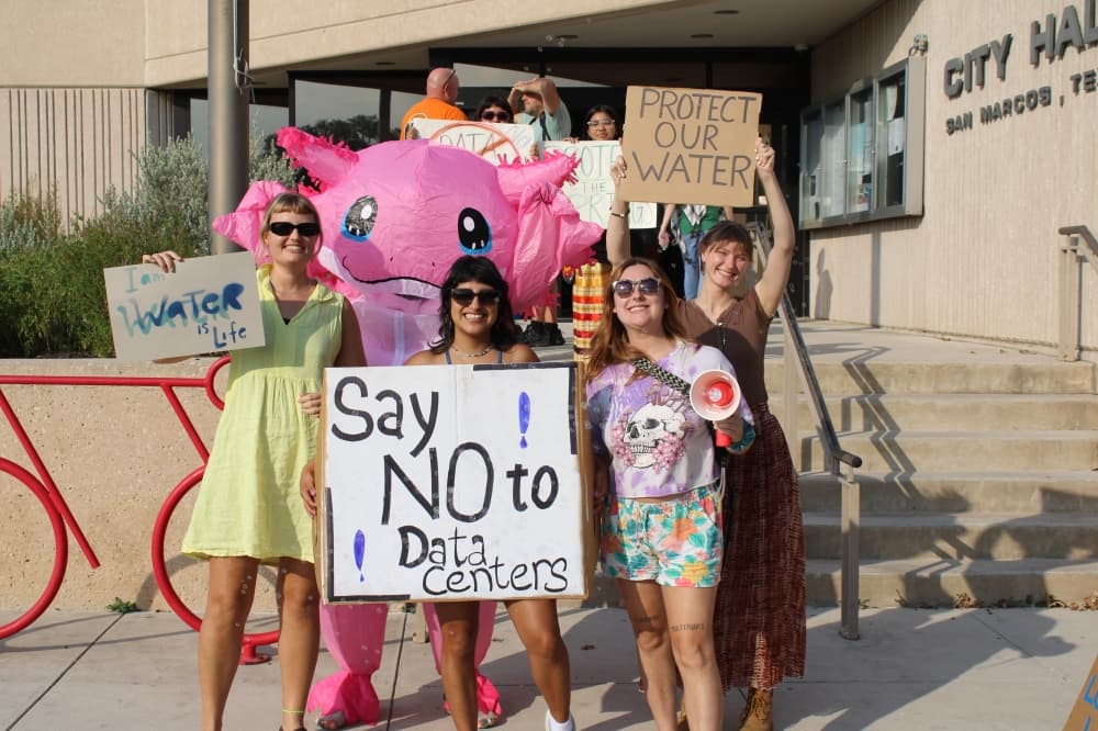 A group of residents in bright colored clothing hold hand-made signs in opposition of the data center. A white sign in the center of the group reads "Say NO to data center." A person in a large inflatable pink axolotl costume stands in the back.