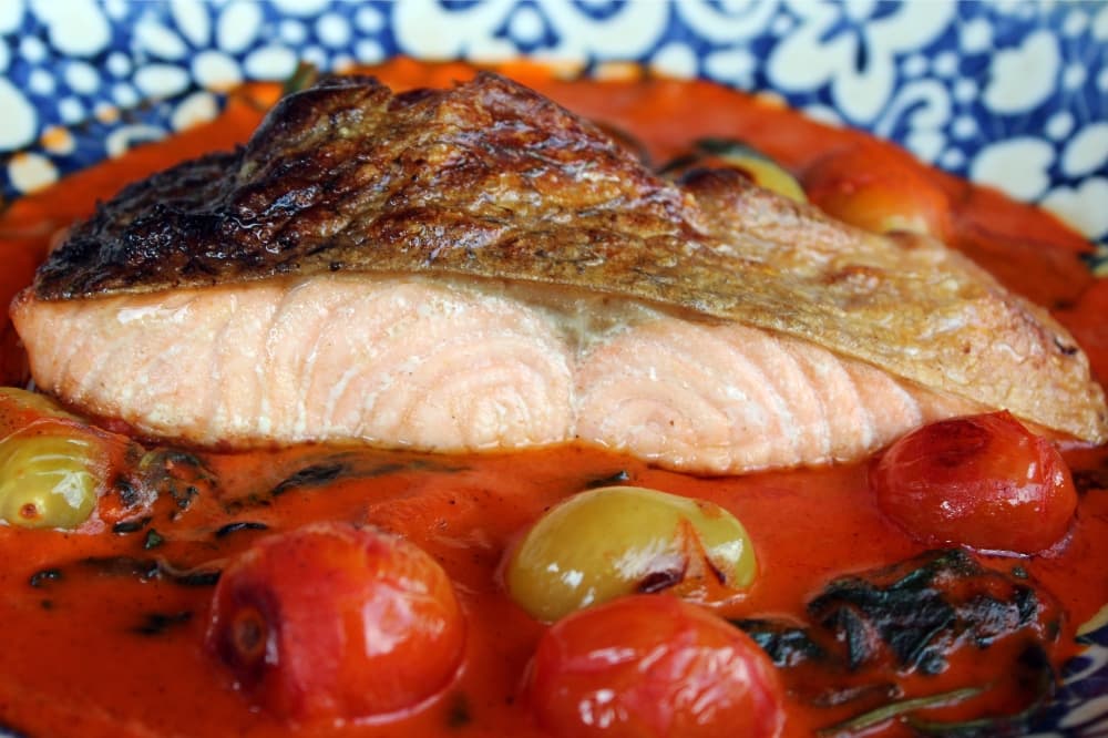 Piece of salmon on a red sauce surrounded by various tomatoes. 