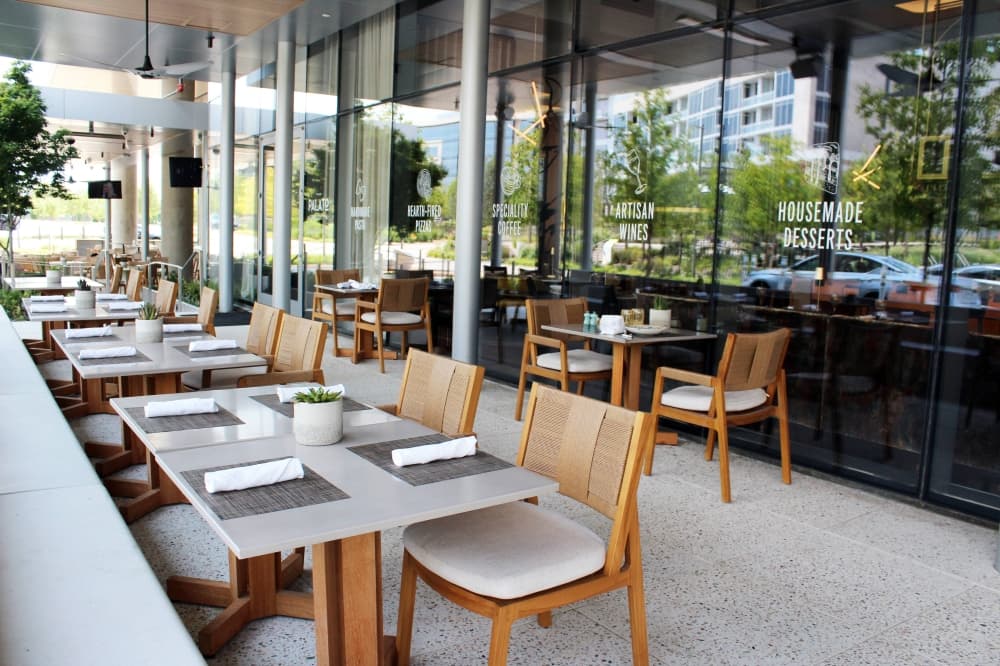 Restaurant patio seating. Floor to ceiling windows reflect green space and office buildings. 