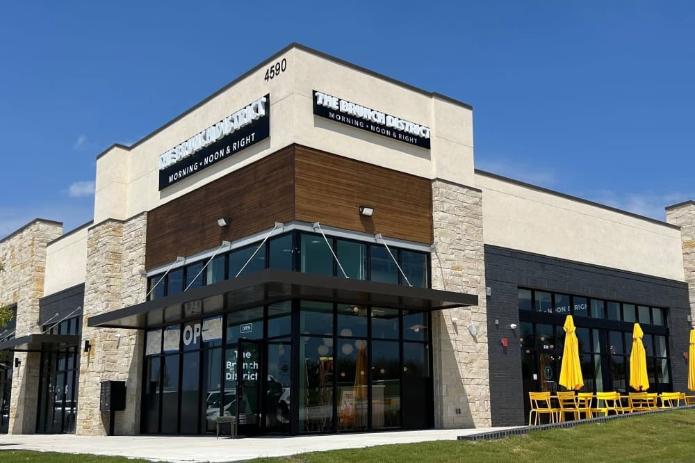 Exterior of corner spot in a strip mall with The Brunch District signage and yellow patio umbrellas by tables. 