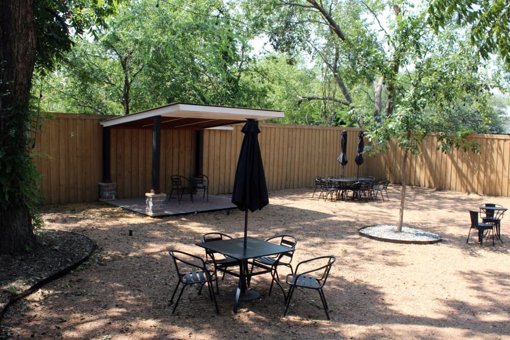 Patio tables, chairs and umbrellas in a fenced in yard with tall trees. 