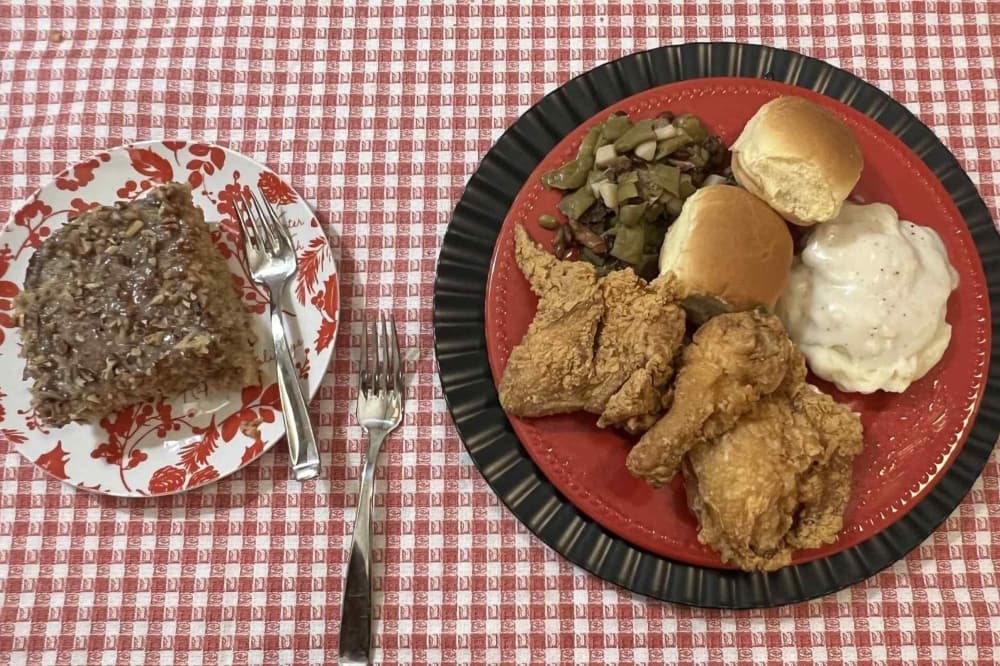 A red and white tablecloth holds a plate of fried chicken with greens and mashed potatoes and rolls. A plate is nearby with a dessert.