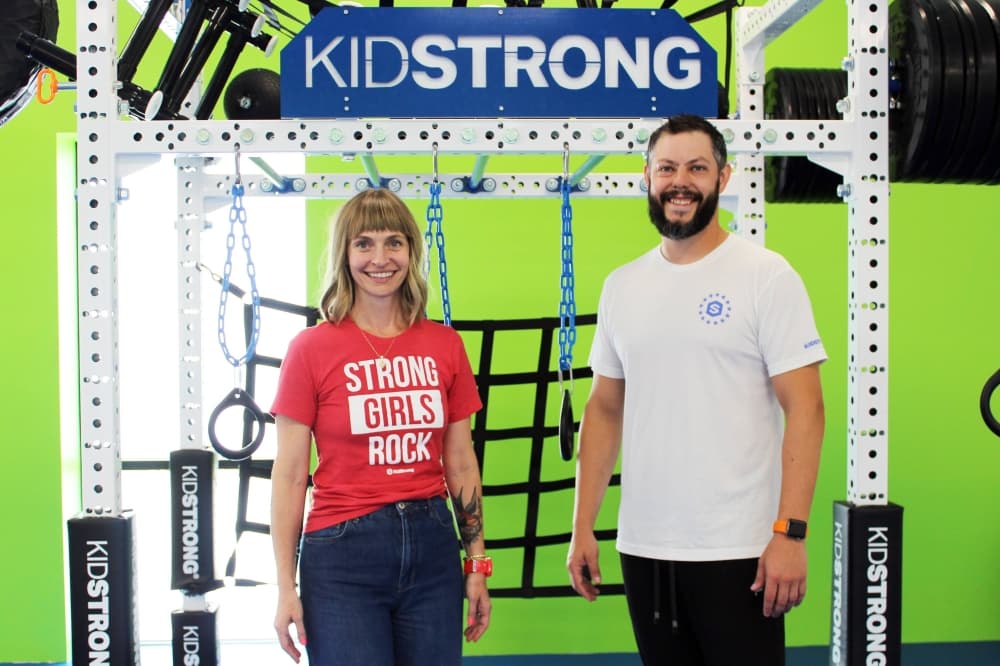 A female and male standing in front of a work out apparatus featuring KidStrong branding. . 