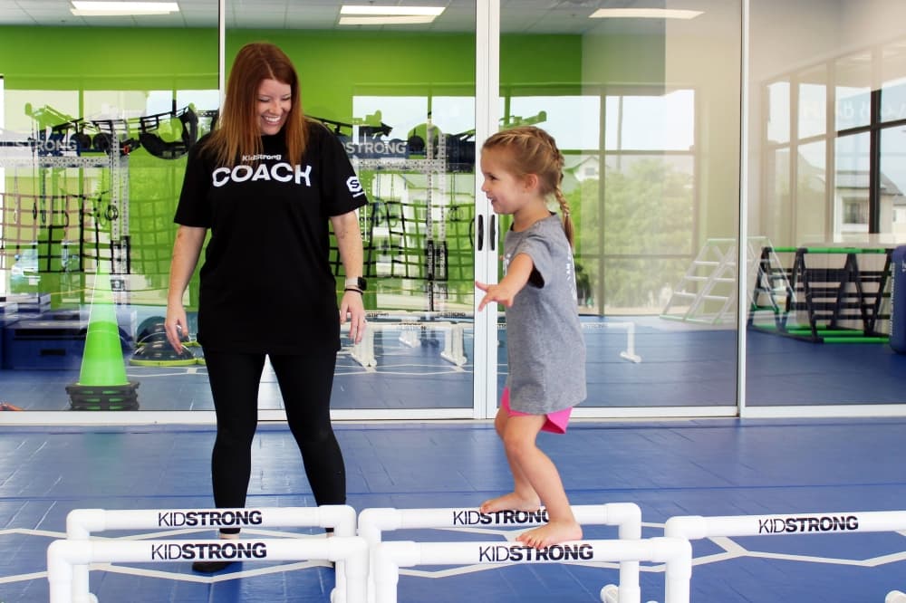 One girl balancing on beams while a woman wearing a coach shirt smiles while watching. 