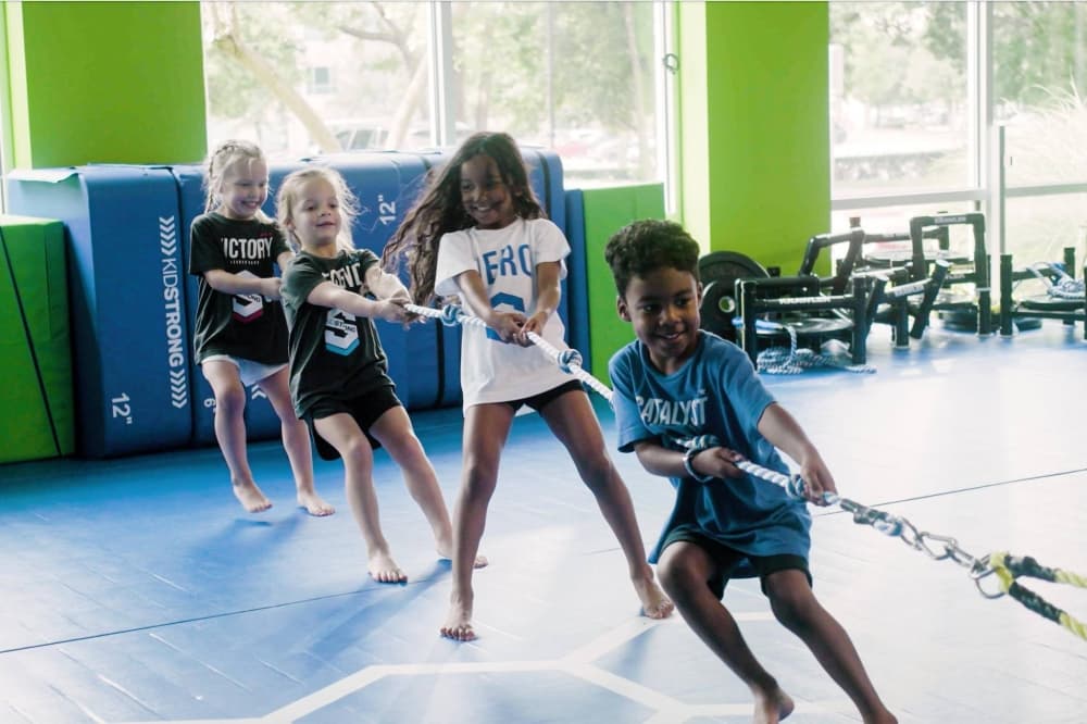 Four kids playing tug-of-war on a blue mat inside a gym-setting. 