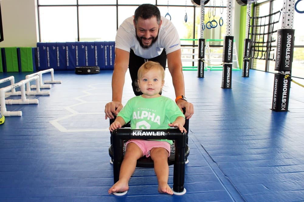 An adult male pushes a baby girl in a workout sled. 