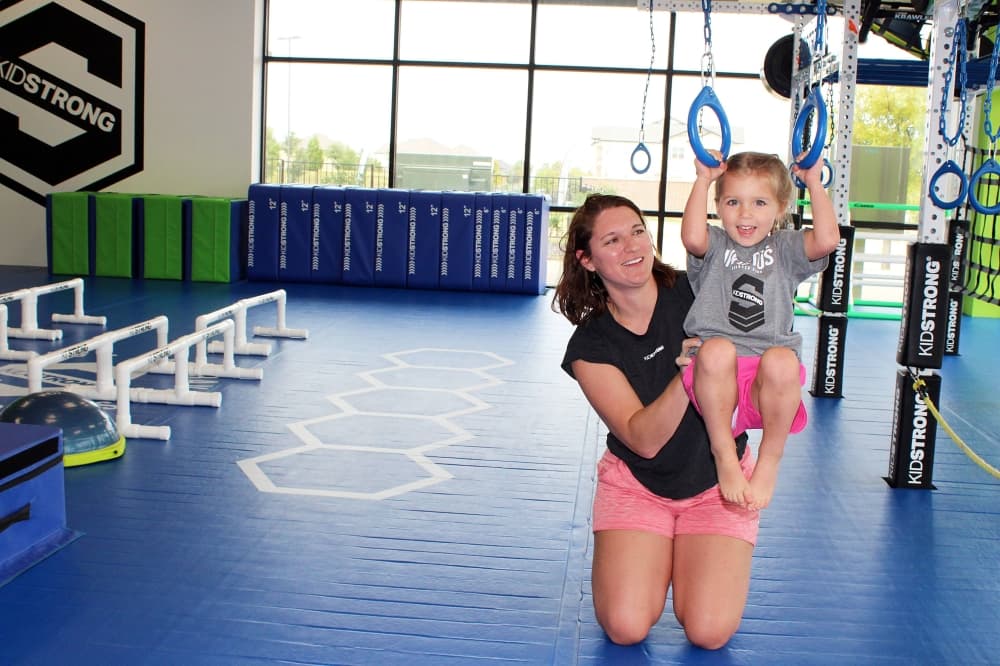 Adult female holds waist of young girl who is holding exercise rings. 