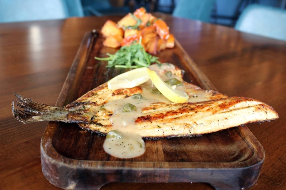 Filet of fish topped with sauce and capers on a wooden tray with sides of vegetables.