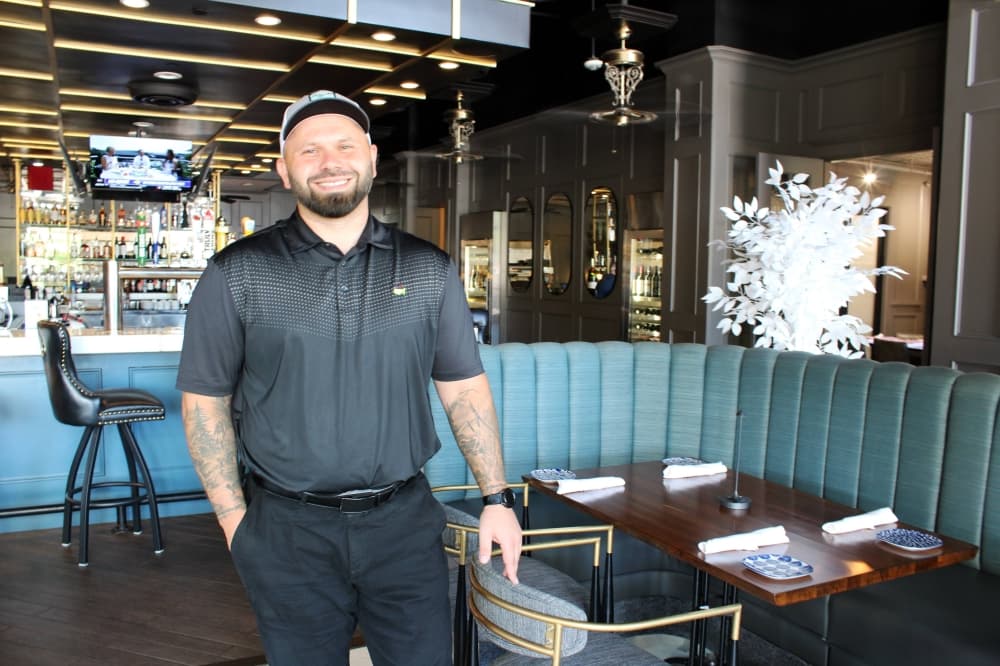 Male standing next to a table in a restaurant with a bar in the background. 