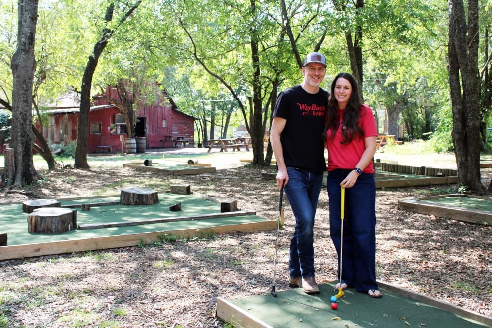 Male and female standing next to each other on a mini golf course surrounded by trees. 