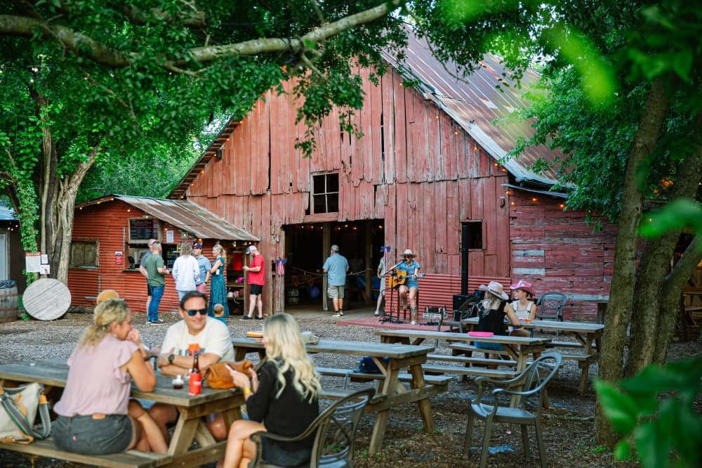 Old red barn with a guitarist performing in the background and groups of people sitting at picnic tables in the foreground. 