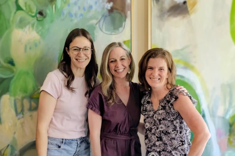 Three females standing side by side in front of a wall. 