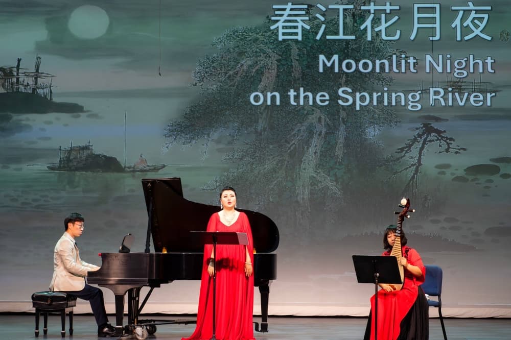 Female wearing a red dress sings on stage between a pianist and a person playing s stringed instrument. 