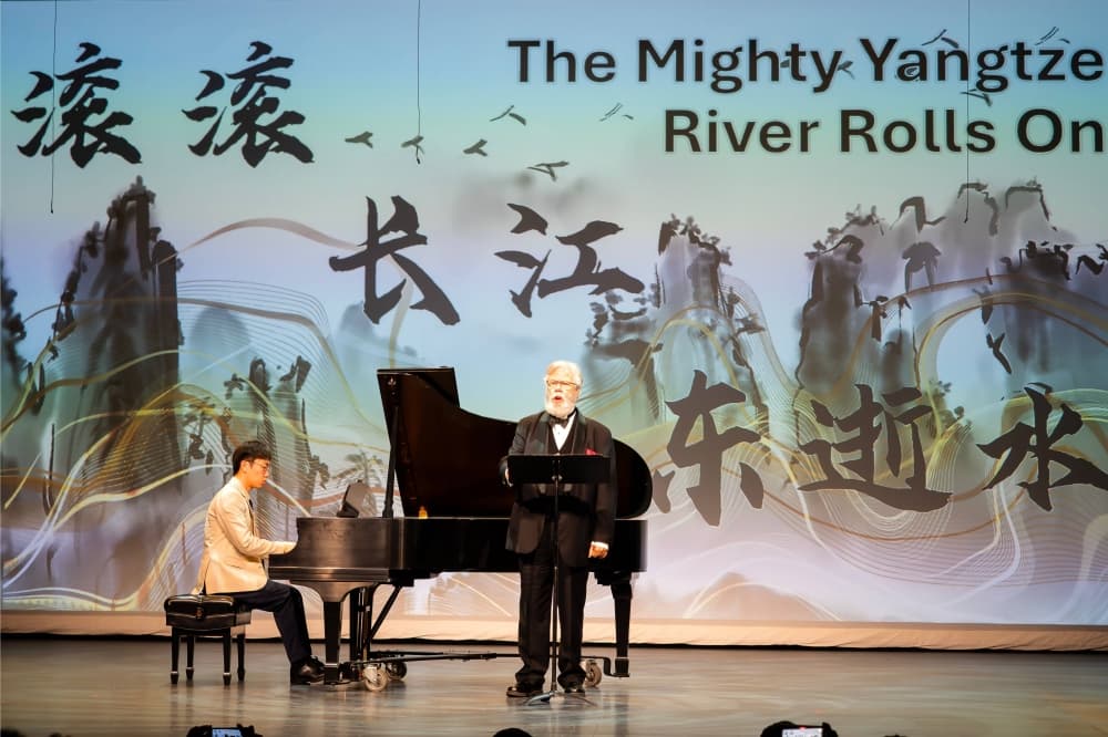 Male wearing a tuxedo sings next to a piano and pianist on a stage.