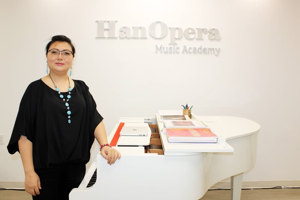 Female standing next to a white baby grand piano in front of a wall featuring HanOpera Music Academy signage. 