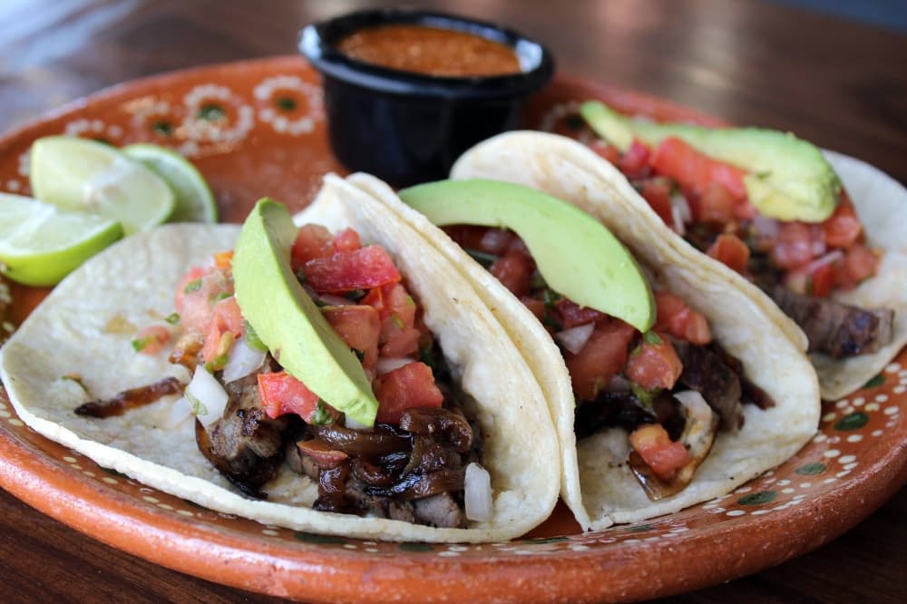 Three steak tacos in soft flour shells topped with pico de gallo and avocado. 