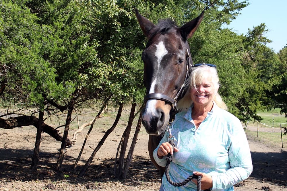 Female standing next to a horse in a field with trees in the background. 