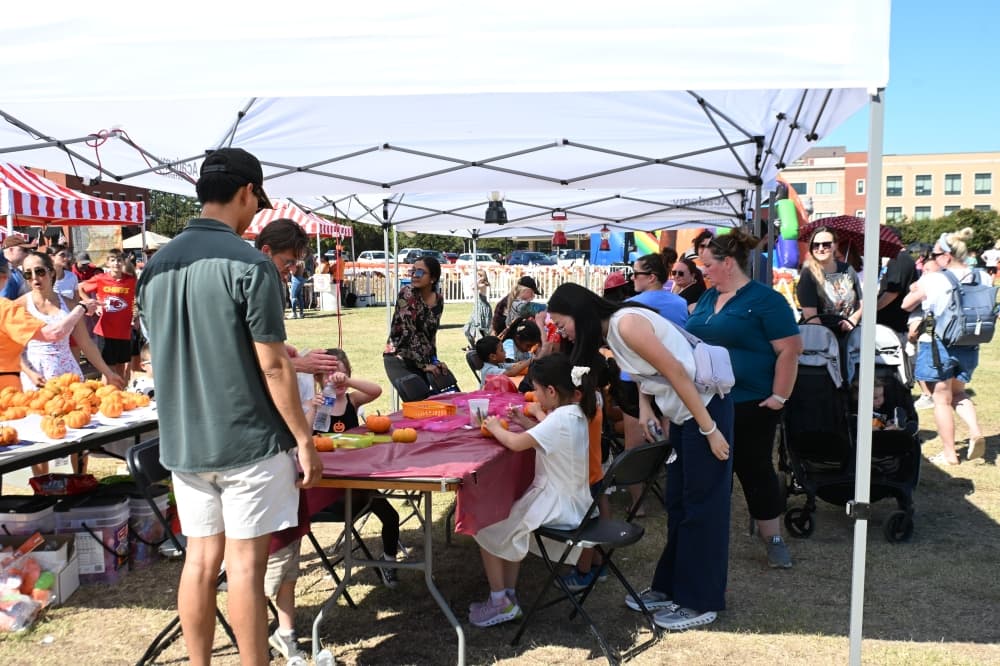 kids decorate pumpkins at a table under a white tent as adults look on