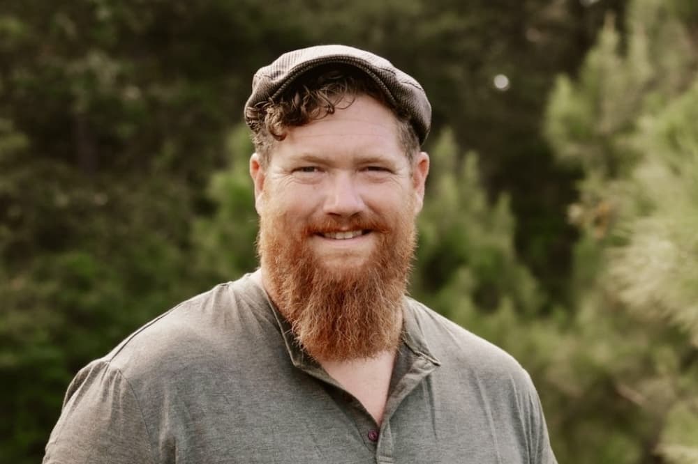 man with a beard smiles in front of a tree background