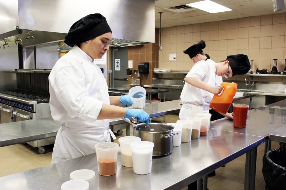 Two males ,wearing chef hats and coats, mixing ingredients in an industrial kitchen. 