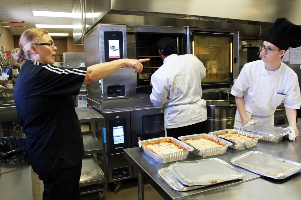 Female wearing black shirt and pants points while male teen wearing chef hat and coat follows directions. 