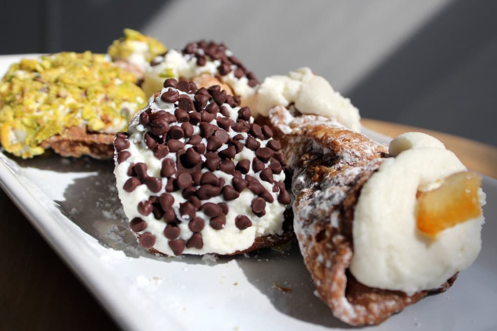 Three cannoli featuring cream filling topped with a variety of items including pistachio, chocolate chips and candied orange slice. 