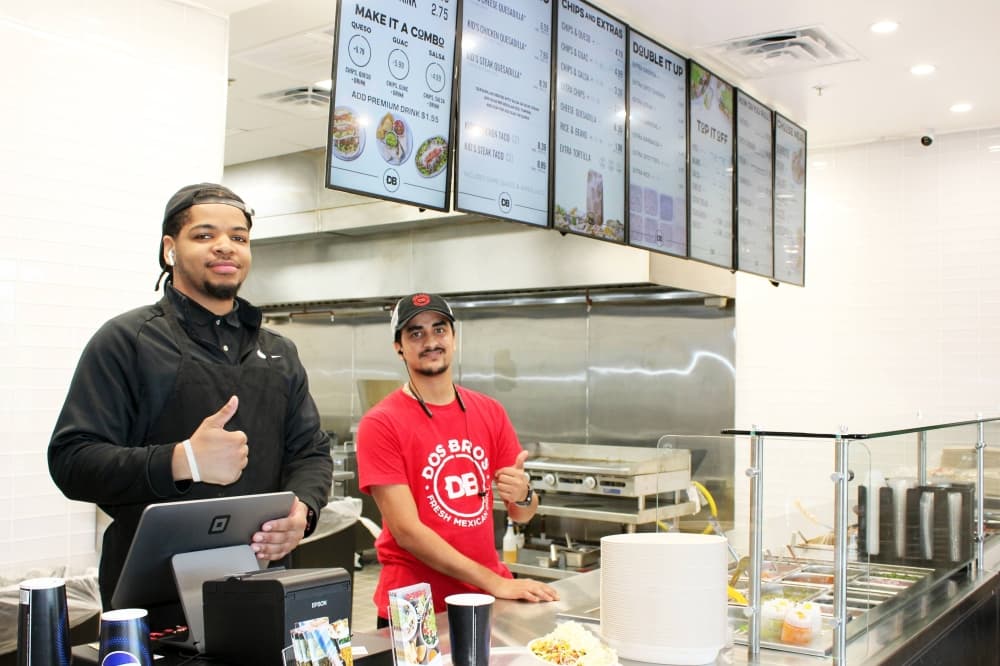 Two males standing behind a counter in an eatery. 