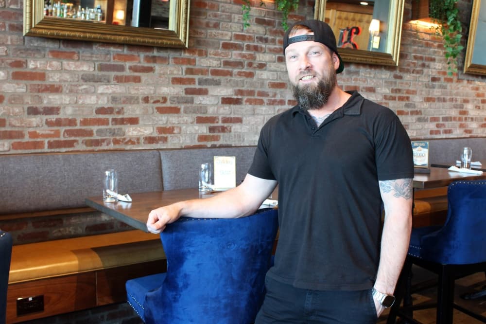 Male wearing a black baseball cap backwards while standing next to a tall chair and table by a brick wall. 