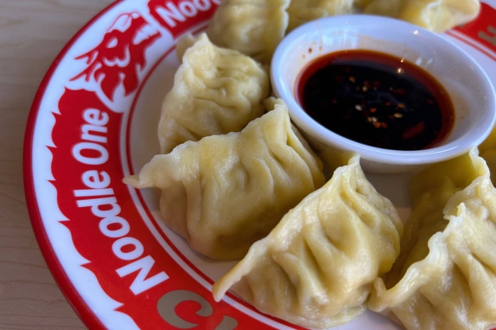 Steamed dumplings and dipping sauce on a dish featuring Noodle One branding. 