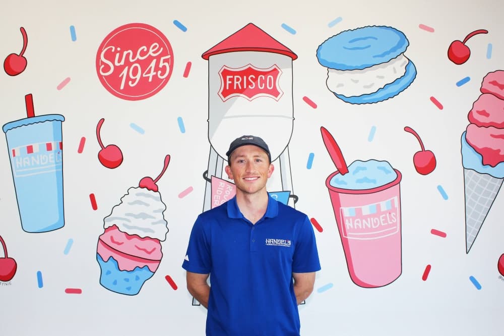 Male wearing a Handel's branded blue shirt standing in front of a indoor mural featuring a water tower with Frisco branding and a variety of ice cream and drinks. 
