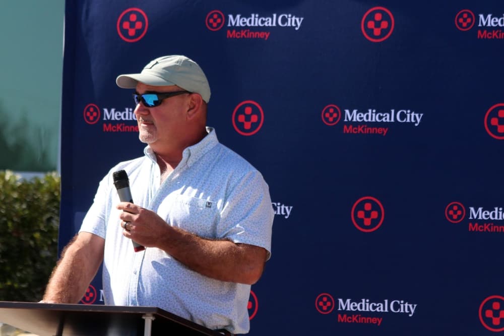 Male holding a microphone standing at a podium in front of a photo screen featuring Medical City of McKinney branding. 
