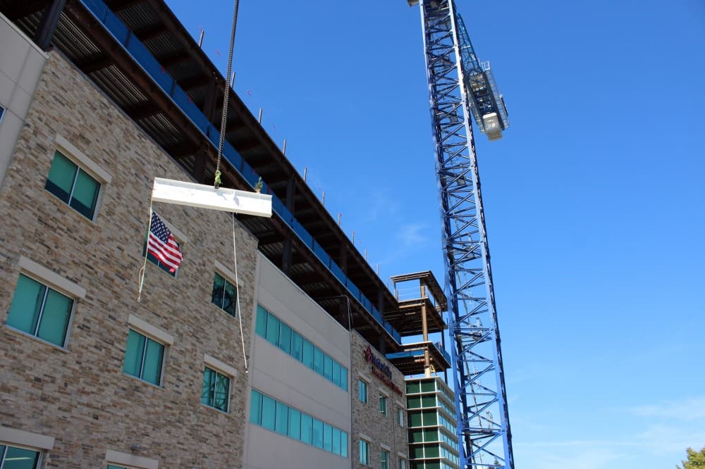 Exterior of a building with a crane hoisting a beam featuring a flag and small tree. 