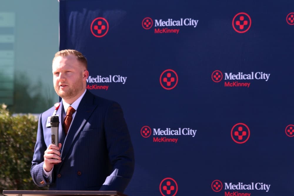 Male holding a microphone standing at a podium in front of a photo screen featuring Medical City of McKinney branding. 