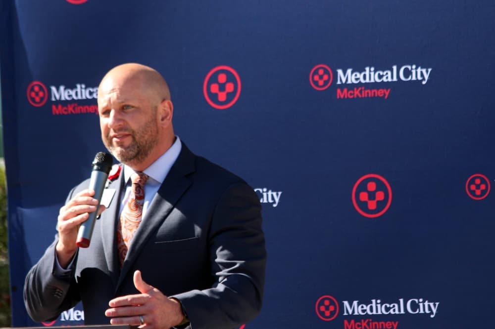 Male holding a microphone standing at a podium in front of a photo screen featuring Medical City of McKinney branding. 