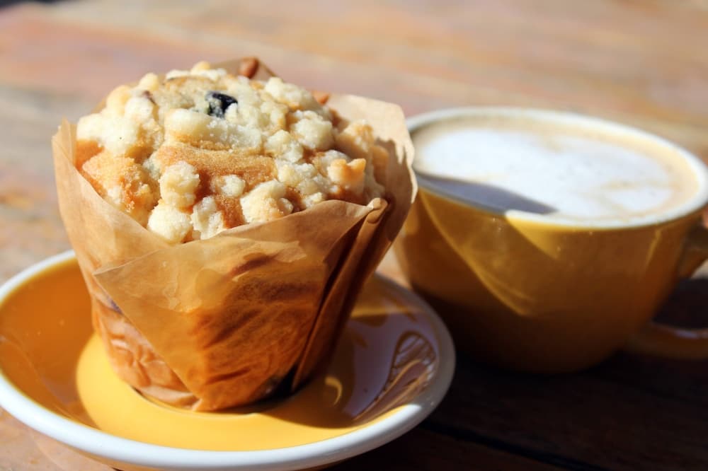 Blueberry muffin on a plate next to a hot latte in a yellow cup. 