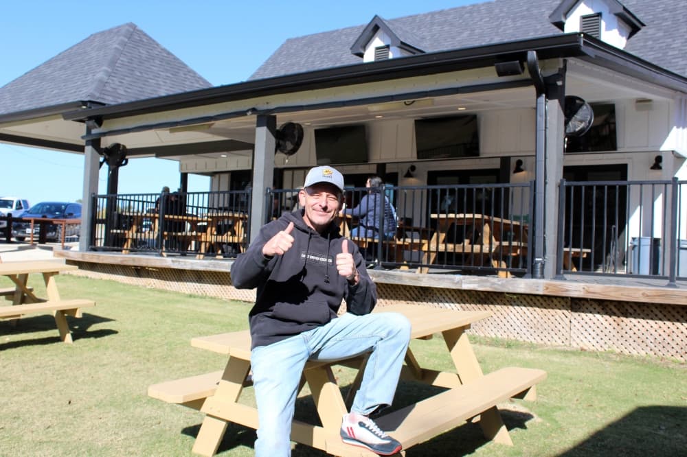 Male leaning on a picnic table holding two thumbs up and smiling. 