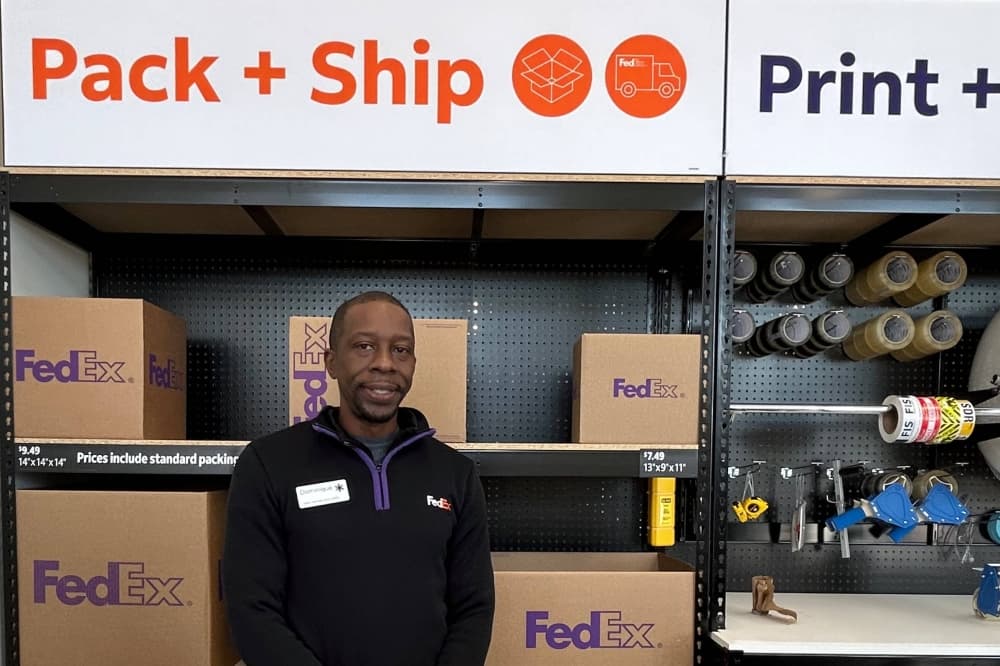 Male wearing FedEx branded quarter-zip sweatshirt standing in front of varying sizes of FedEx boxes in a FedEx Office store. 