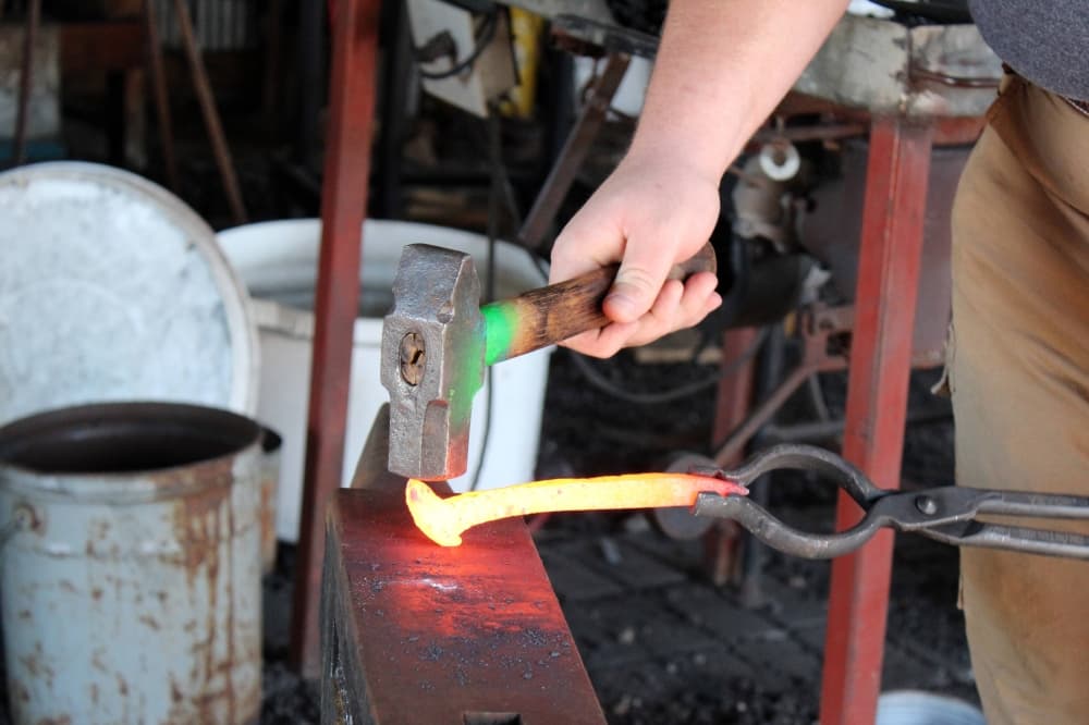 Hand holding a hammer striking a piece of hot metal held by tongs. 
