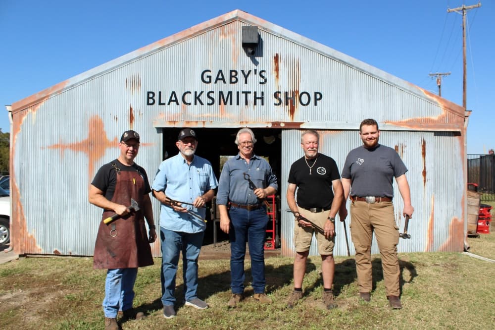 A group of five men holding blacksmith tools standing in front of a building featuring Gaby's Blacksmtih Shop signage. 